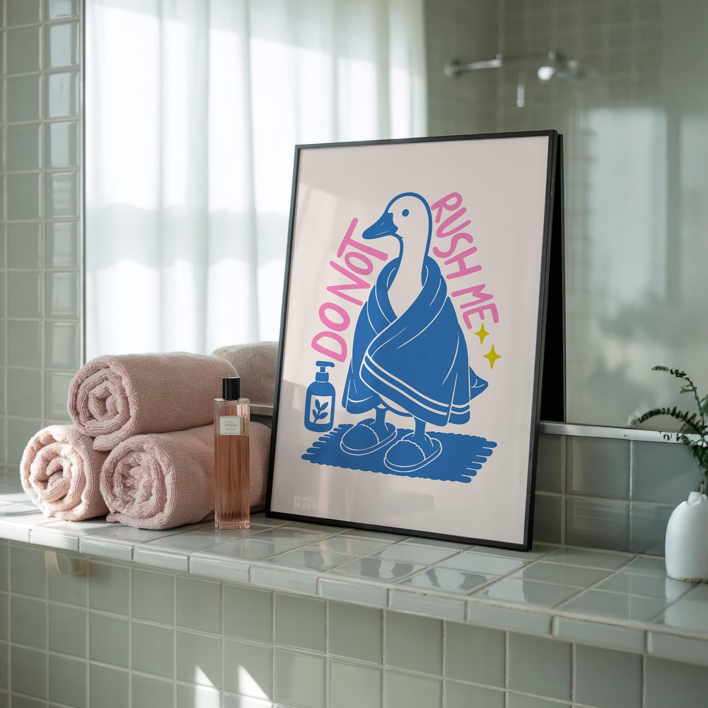 A framed print of a goose in a blue towel and slippers, with "DO NOT RUSH ME" text, rests on a tiled shelf next to rolled pink towels and a perfume bottle.