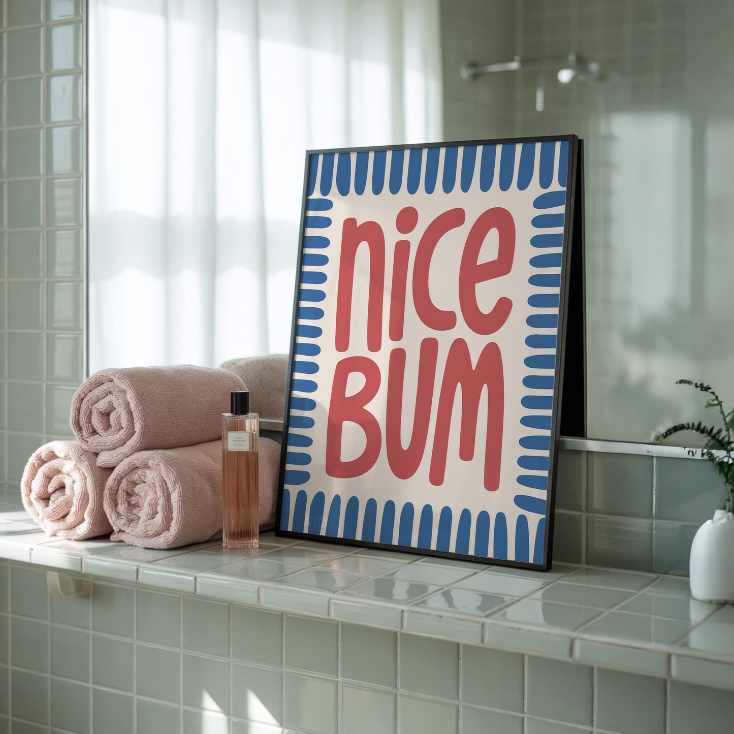 A framed print with "nice BUM" in red, stylized text on a cream background with a blue border, rests on a tiled bathroom ledge next to rolled pink towels.