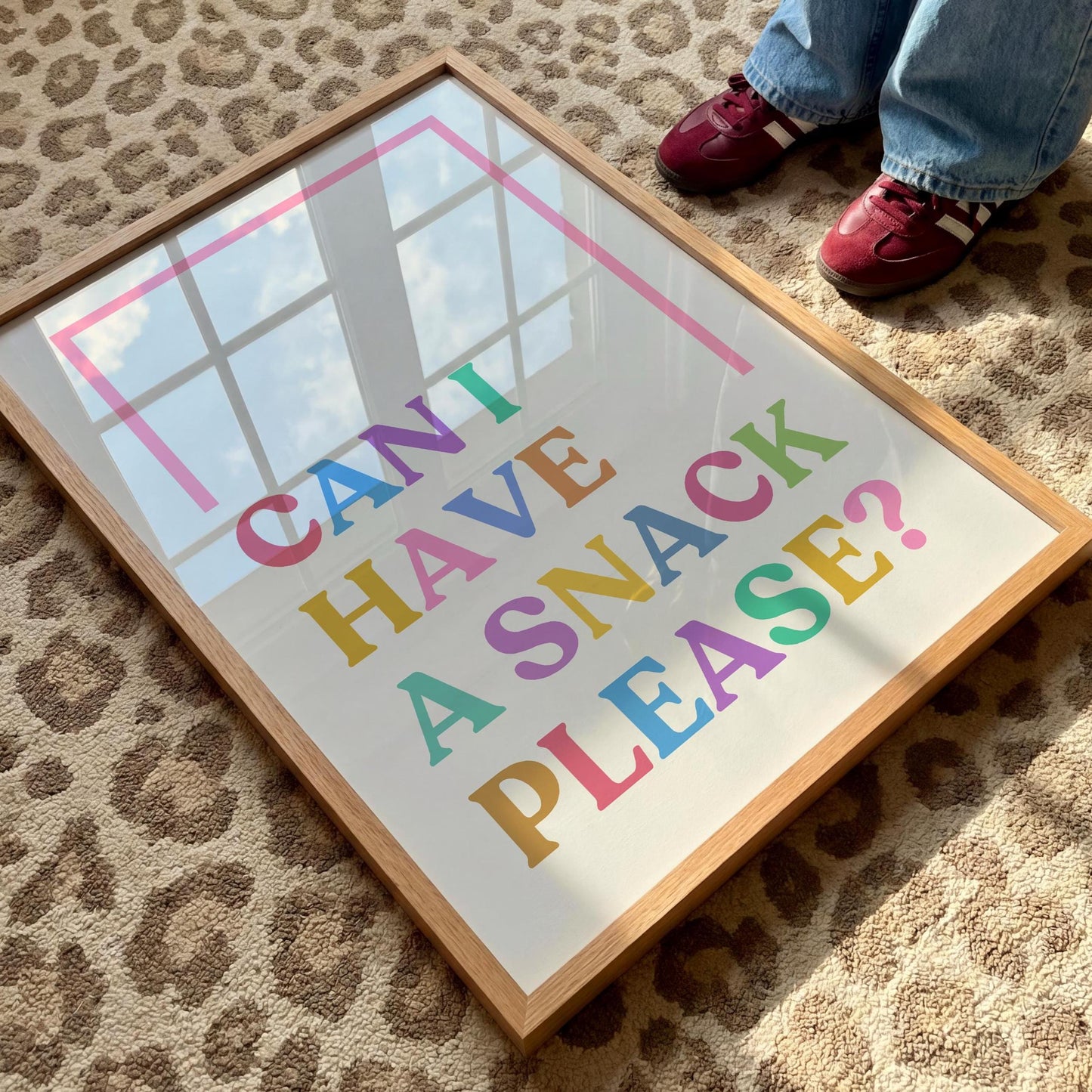 A framed print with "CAN I HAVE A SNACK PLEASE?" in colorful letters, lying on a leopard print rug with a person's feet visible.