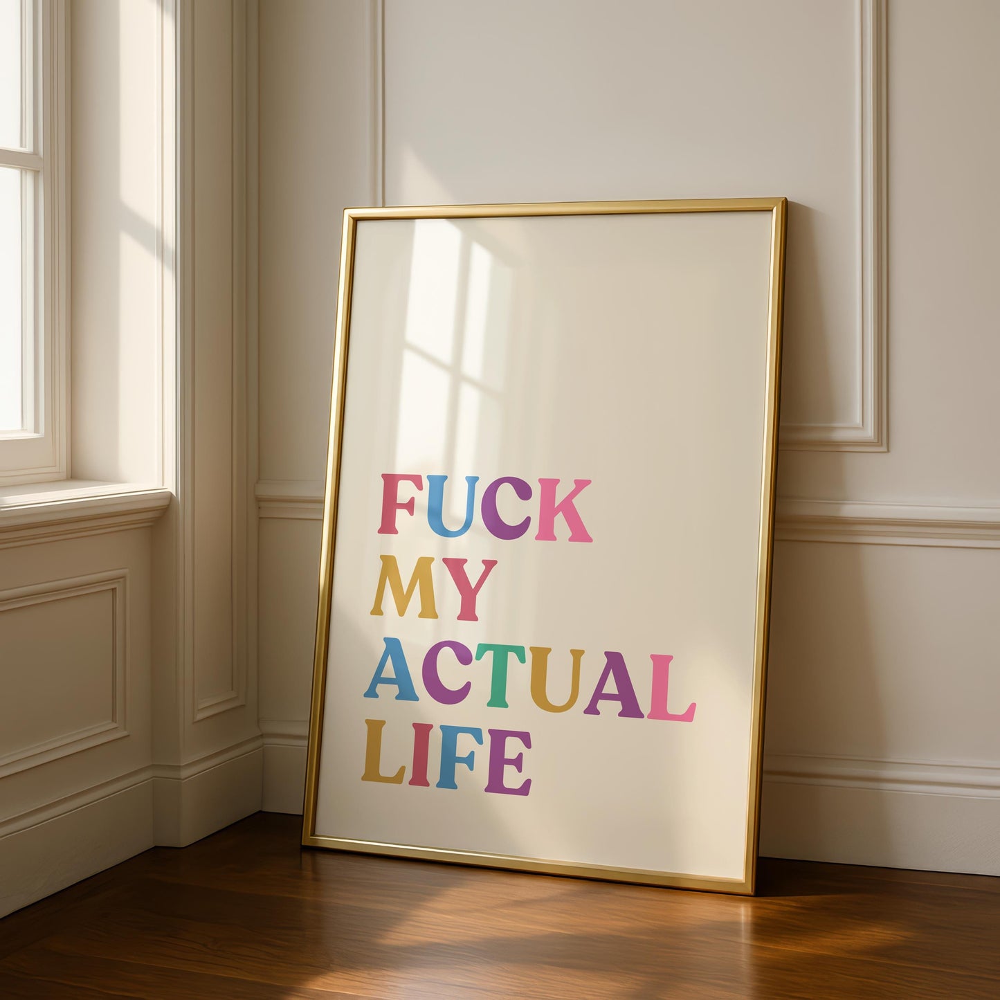 A gold-framed print on a light wall, reading "FUCK MY ACTUAL LIFE" in colorful, block letters, with sunlight streaming in from a window on the left.