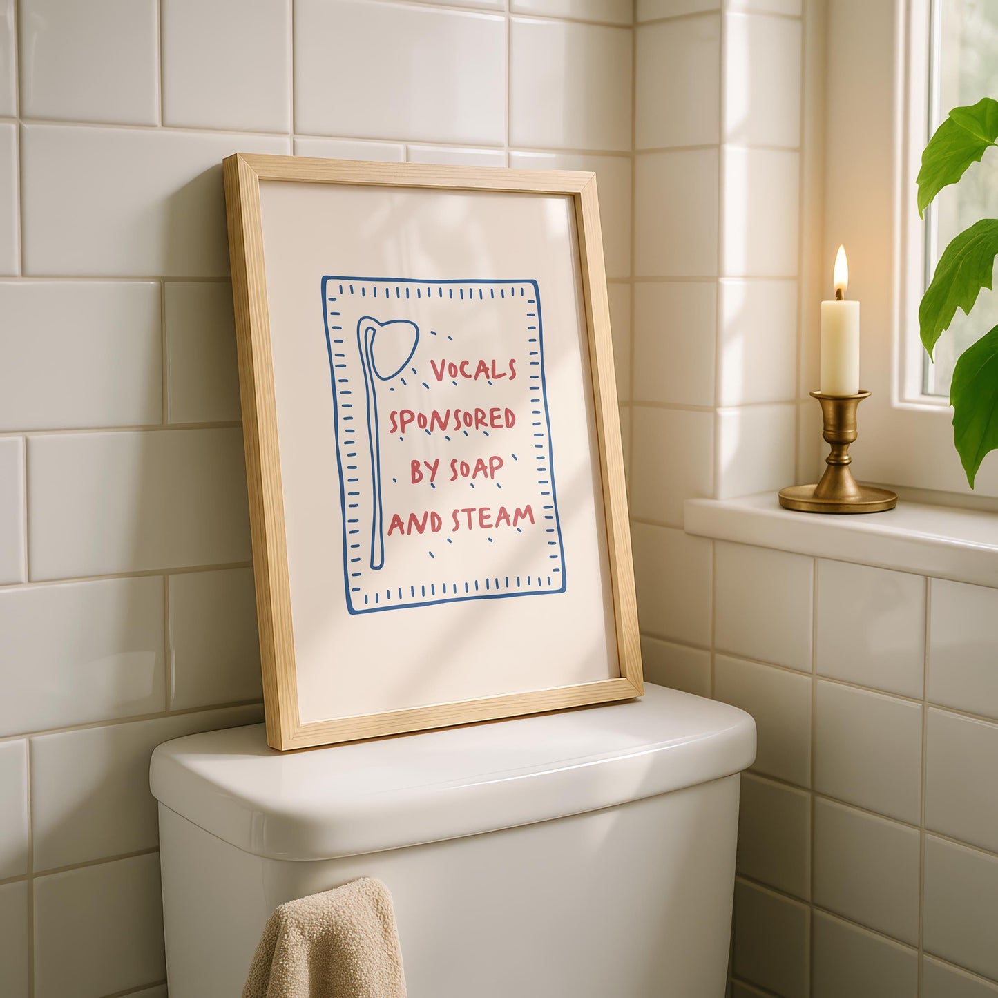 A framed print with a light wood frame leans against a white tiled wall on top of a toilet, displaying a red and blue design on a cream background that reads "VOCALS SPONSORED BY SOAP AND STEAM."