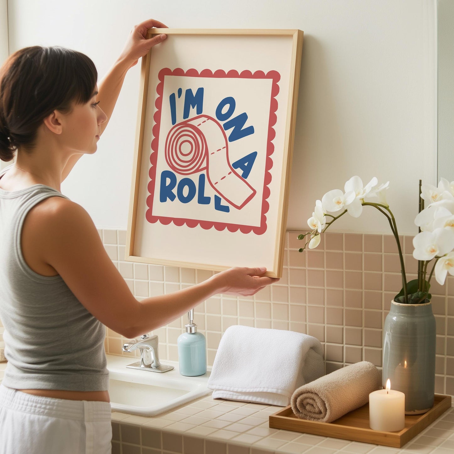 A woman in a gray tank top hangs a light wood-framed print with "I'M ON A ROLL" and a toilet paper roll, above a white sink with a blue soap dispenser and white towels.