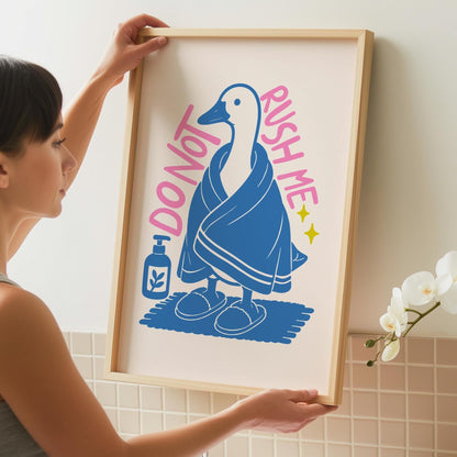 A person holds up a framed print of a white goose in a blue towel and slippers, with text "DO NOT RUSH ME" and a blue soap bottle.
