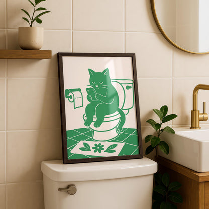 A framed print of a green cat on a toilet looking at a phone, with a toilet paper roll and a green flower mat, sits on a toilet tank.