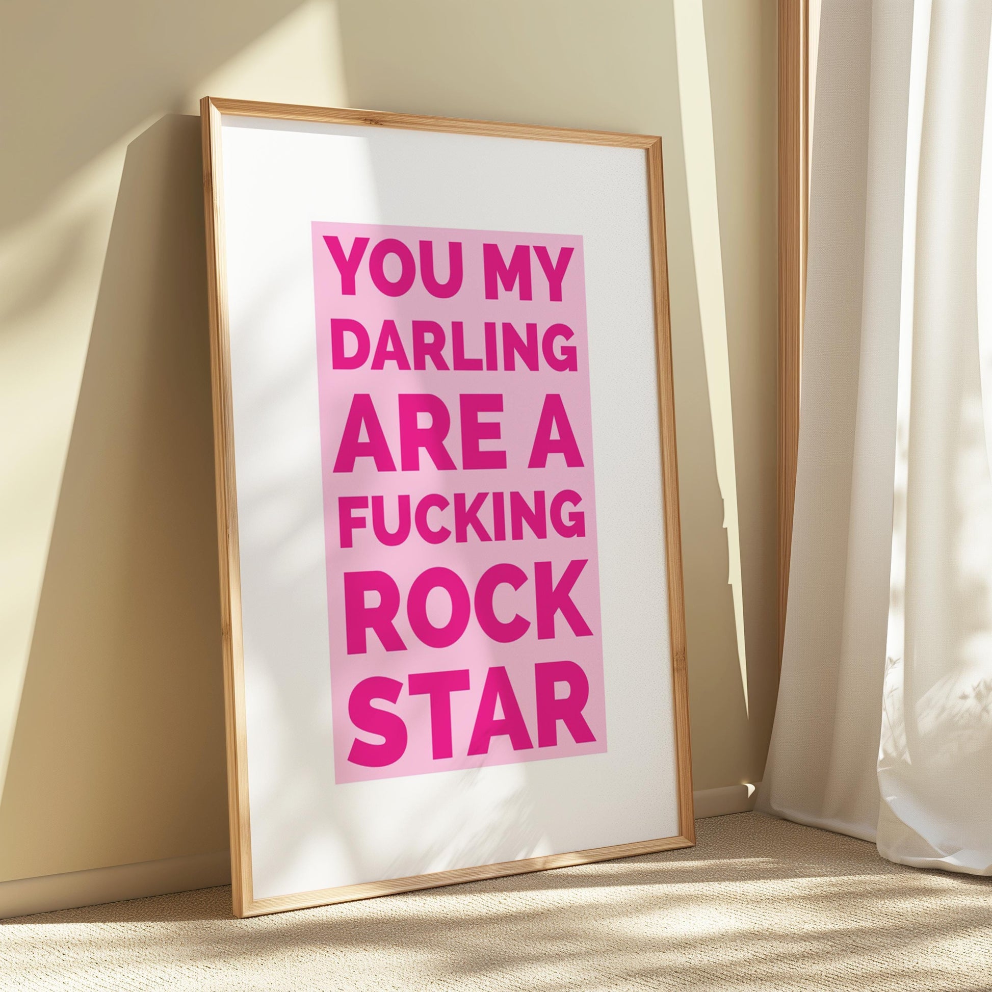 A framed print with a gold border leans against a light brown wall, displaying "YOU MY DARLING ARE A FUCKING ROCK STAR" in pink text on a white background.