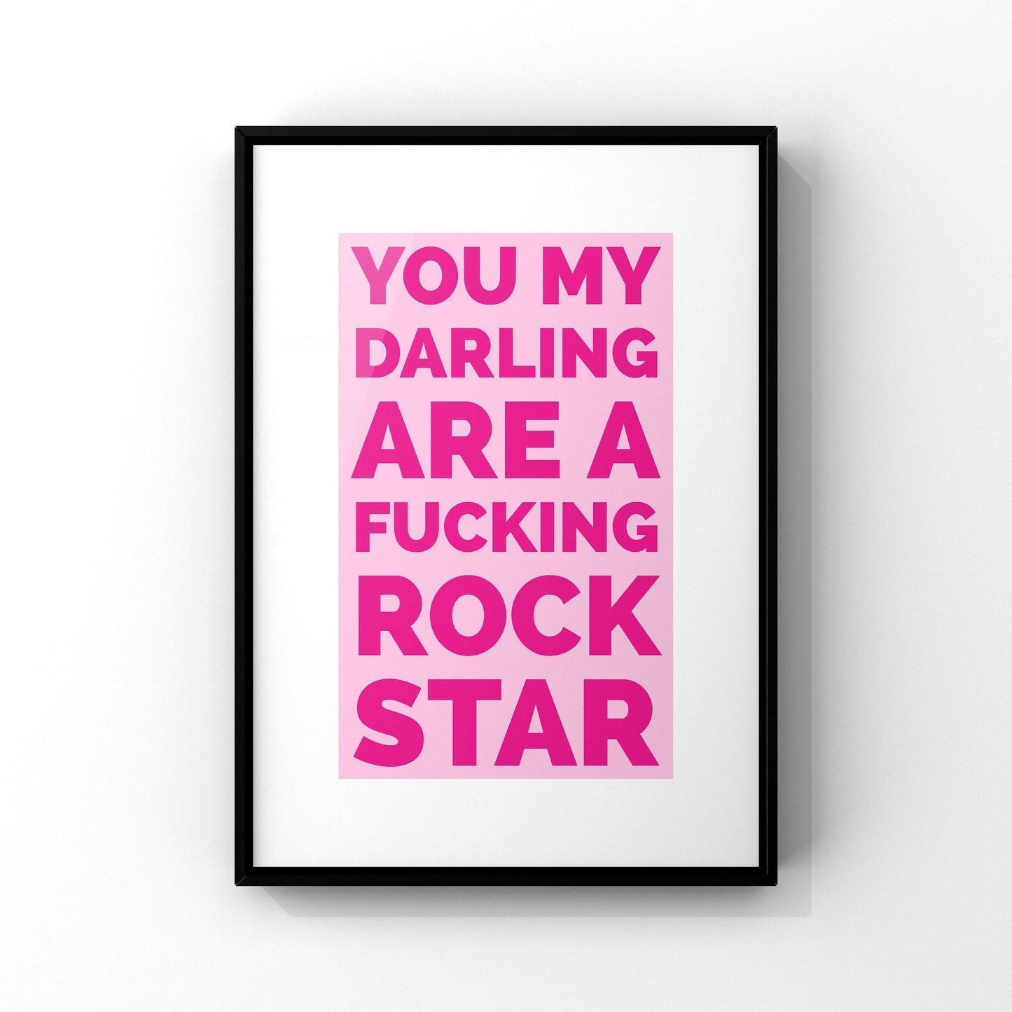 A black-framed print hangs on a white wall, featuring "YOU MY DARLING ARE A FUCKING ROCK STAR" in bold pink letters on a lighter pink background.