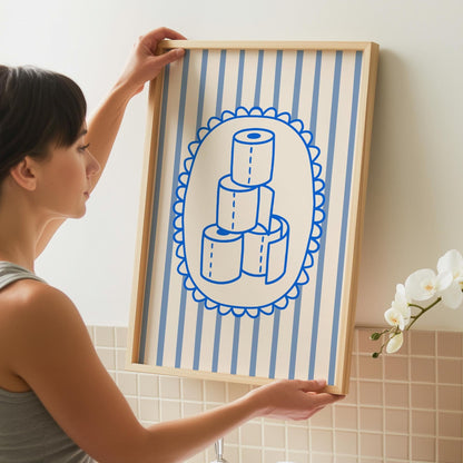 A person holds a framed print with a light wood frame, depicting a stack of four toilet paper rolls in blue outline on a white oval, set against a background of light blue and white vertical stripes.