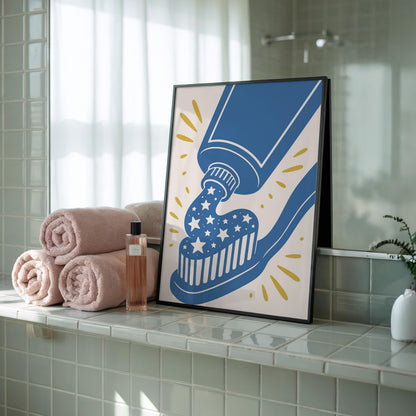 A framed print of a blue toothpaste tube squeezing white toothpaste with blue stars onto a blue toothbrush, against a cream background with yellow starbursts, rests on a tiled bathroom counter next to rolled pink towels and a perfume bottle.