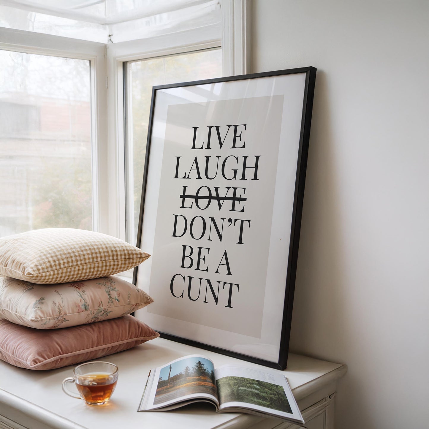 A black-framed print with the text "LIVE LAUGH LOVE DON'T BE A CUNT" in black on a light background, sitting on a white dresser next to a window with a stack of pillows and a teacup.
