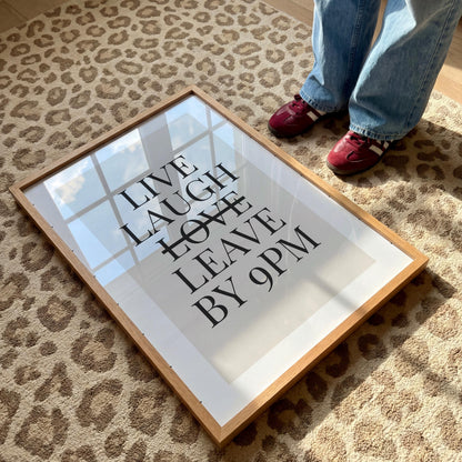 A framed print with a light wood border lies on a leopard print rug, displaying the text "LIVE LAUGH LEAVE BY 9PM" in black sans-serif font, with a horizontal line through "LAUGH."
