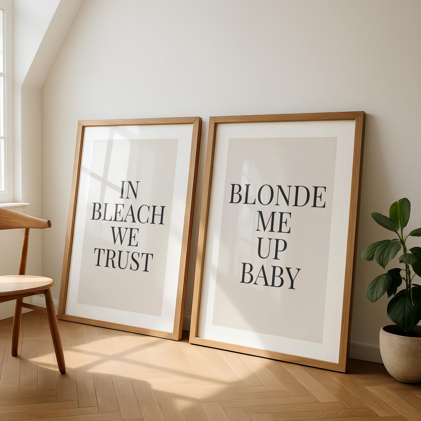 Two framed prints lean against a light wall, one reading "IN BLEACH WE TRUST" and the other "BLONDE ME UP BABY," next to a wooden chair and a potted plant.