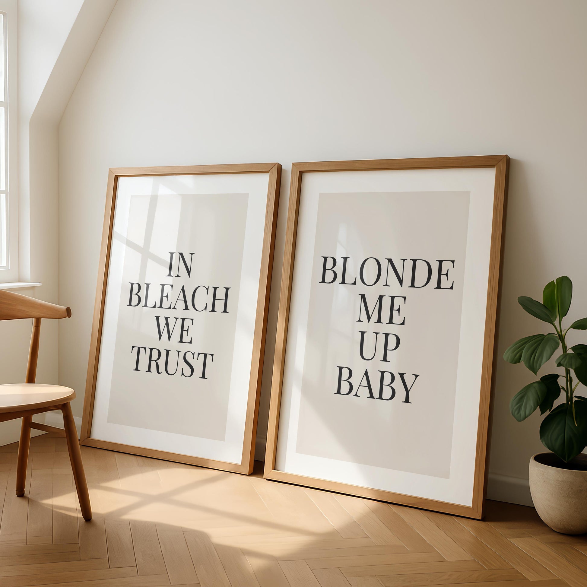 Two framed prints lean against a light wall, one reading "IN BLEACH WE TRUST" and the other "BLONDE ME UP BABY," next to a wooden chair and a potted plant.