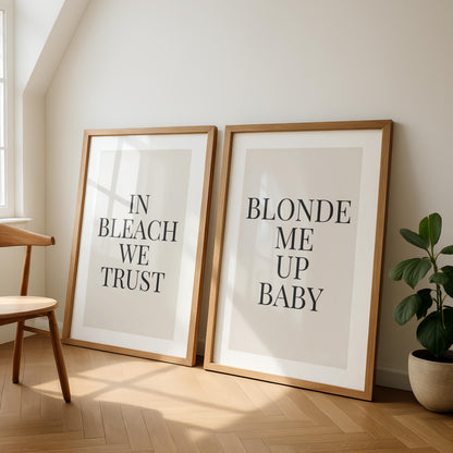 Two framed prints lean against a light wall, one reading "IN BLEACH WE TRUST" and the other "BLONDE ME UP BABY," next to a wooden chair and a potted plant.