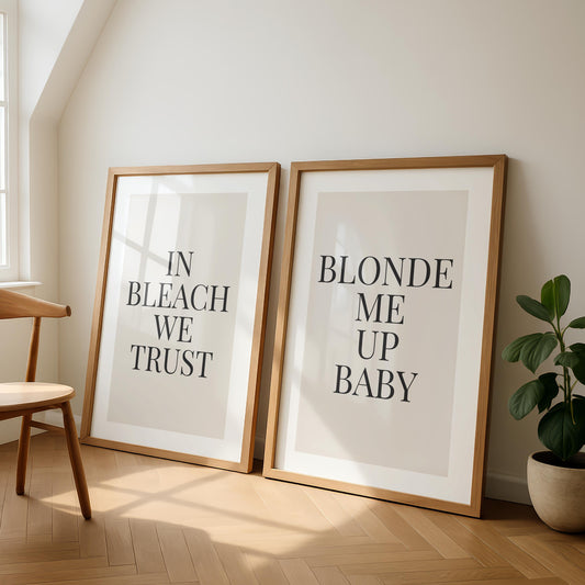 Two framed prints lean against a light wall, one reading "IN BLEACH WE TRUST" and the other "BLONDE ME UP BABY," next to a wooden chair and a potted plant.