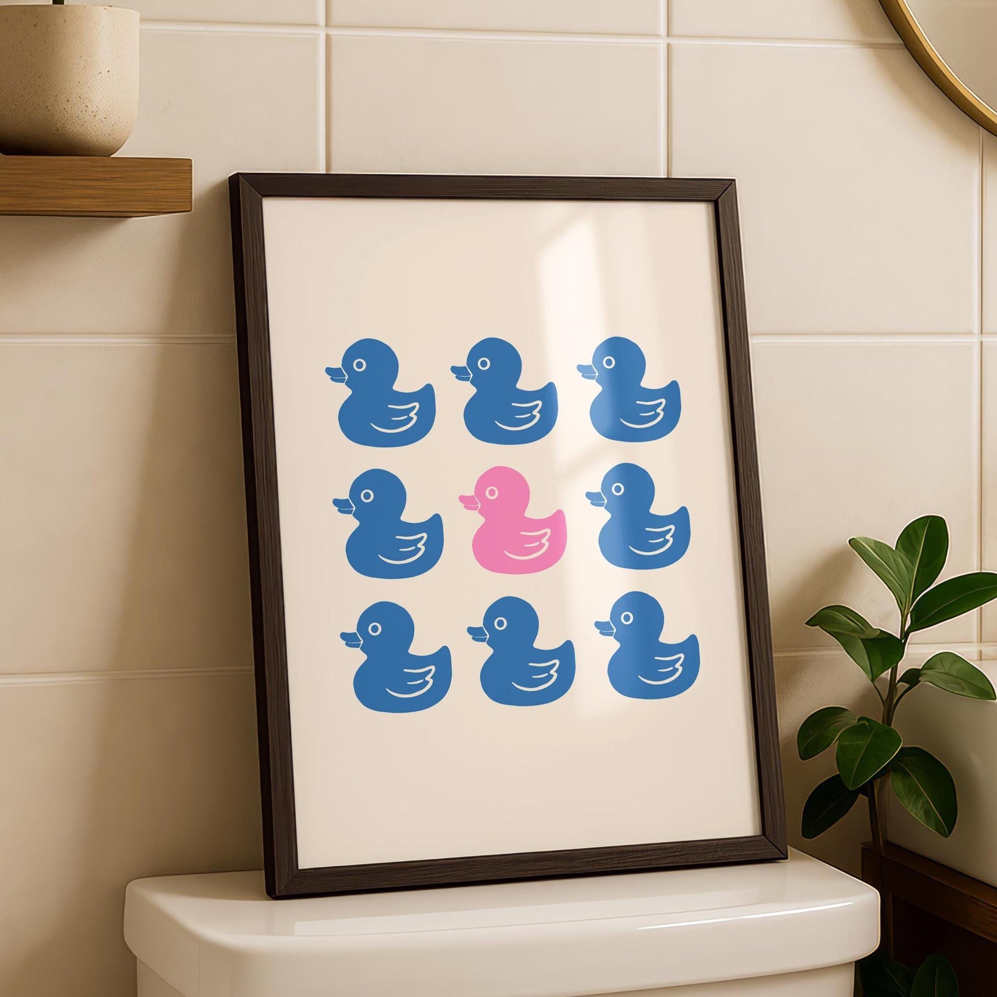 A framed print with nine rubber ducks, eight blue and one pink, hangs on a white tiled bathroom wall above a toilet.