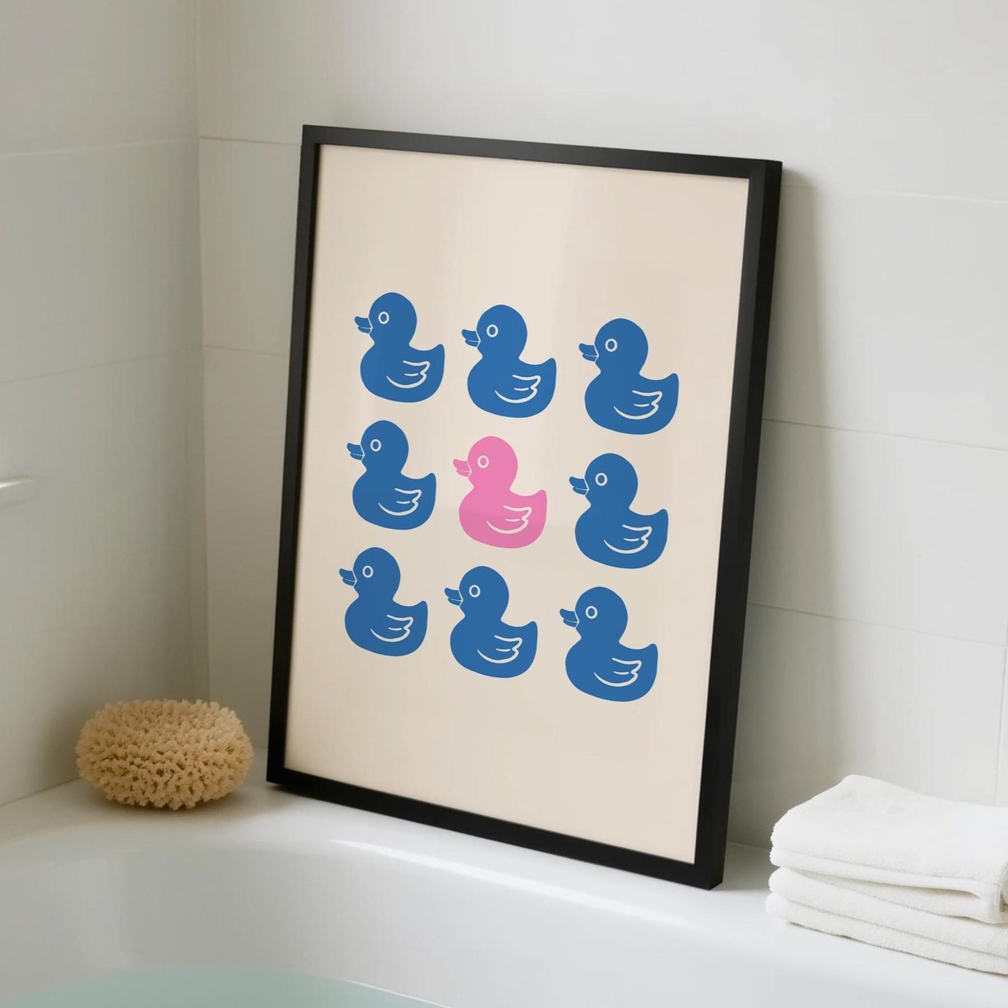 A framed print with nine rubber ducks, eight blue and one pink, leans against a white tiled bathroom wall next to a bathtub with a sponge and towels.