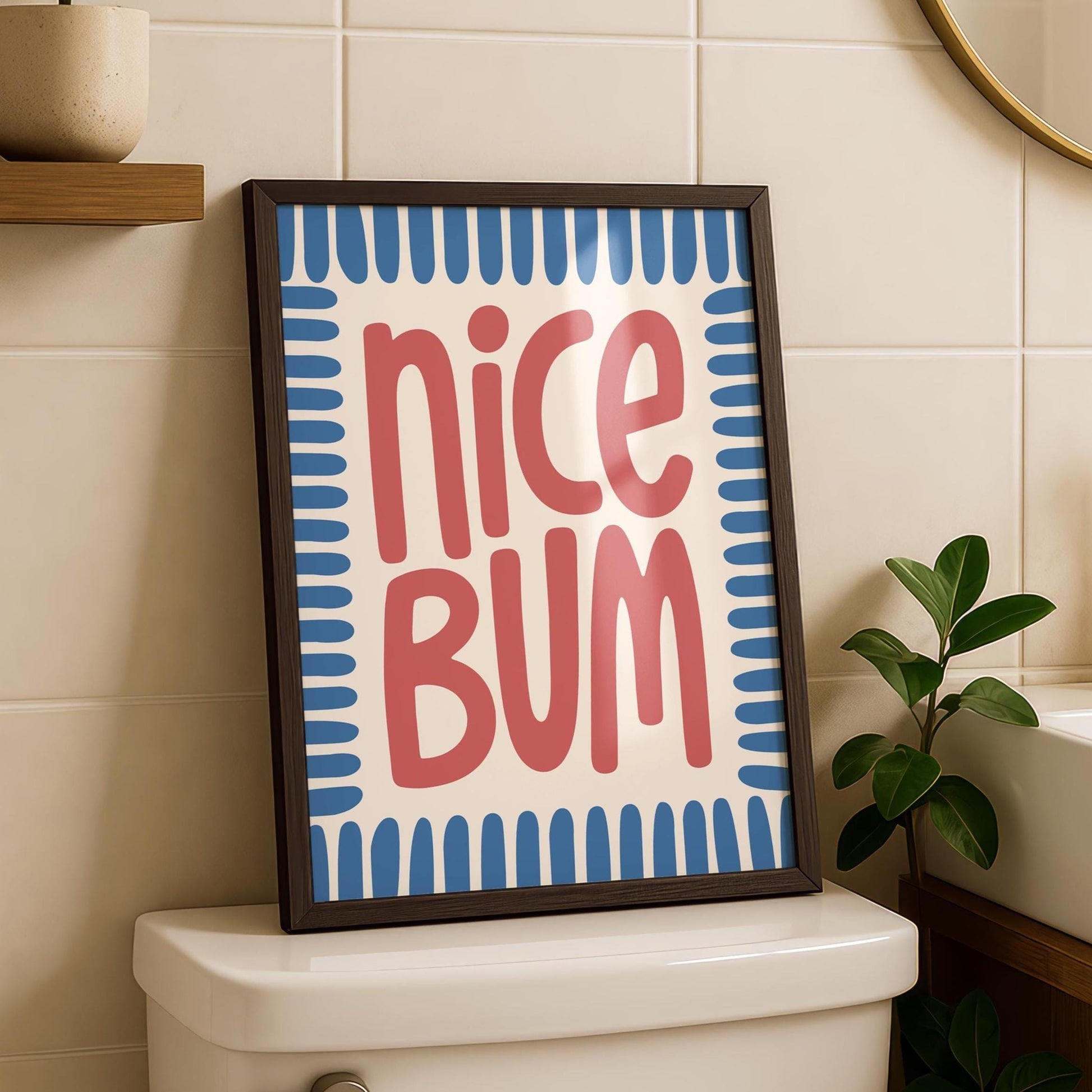 A framed print with "nice BUM" in red, stylized text on a cream background with blue border, sits on a white toilet tank.