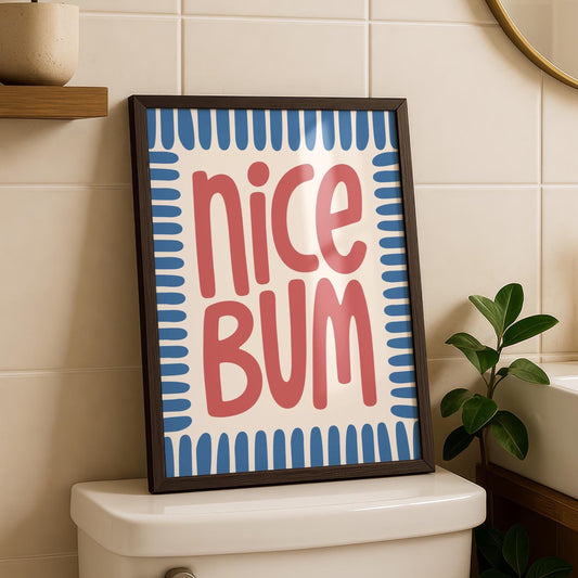 A framed print with "nice BUM" in red, stylized text on a cream background with blue border, sits on a white toilet tank.