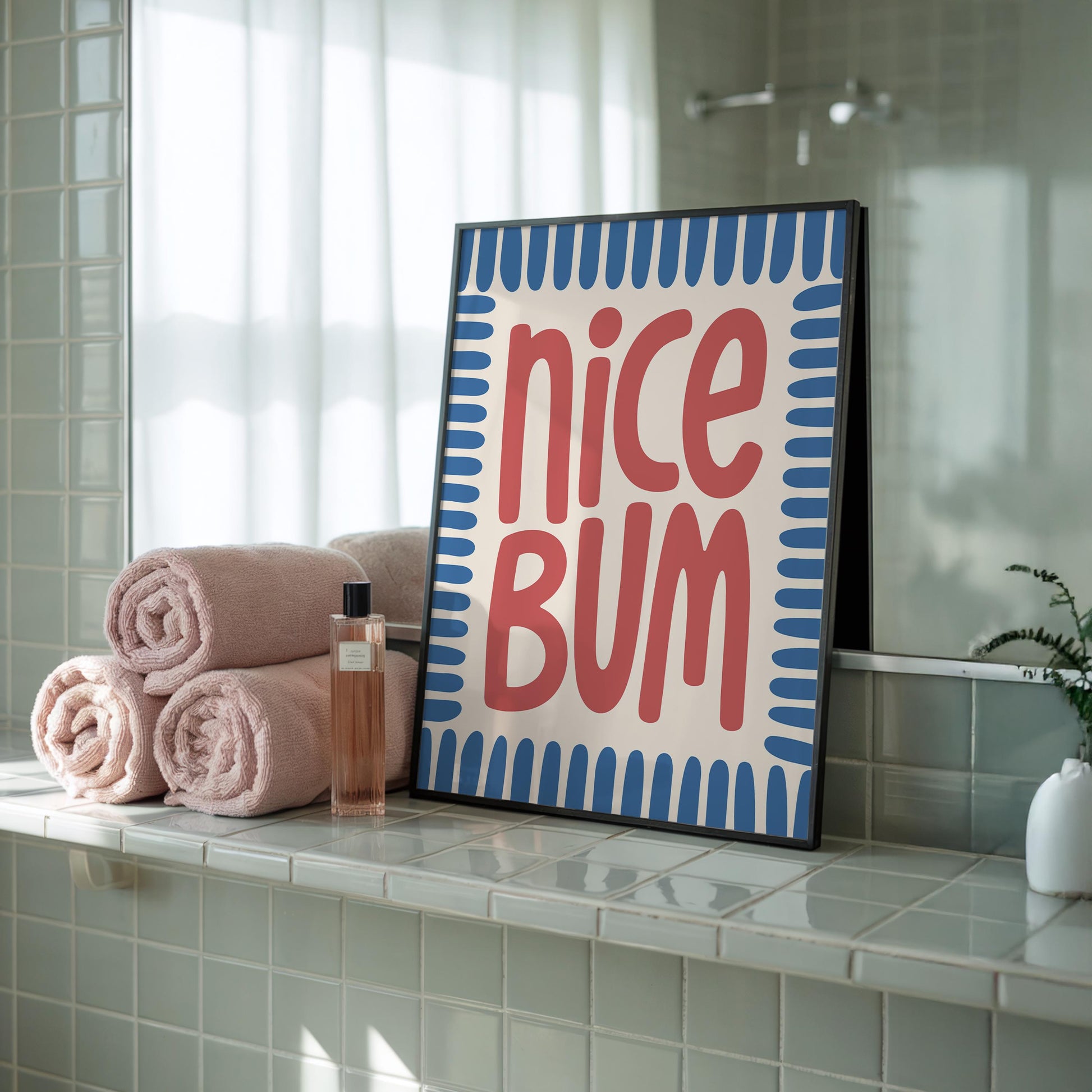 A framed print with "nice BUM" in red, stylized text on a cream background with a blue border, rests on a tiled bathroom ledge next to rolled pink towels.