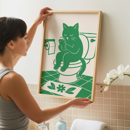 A woman holds up a framed print of a green cat sitting on a toilet and looking at a phone, against a light pink background.