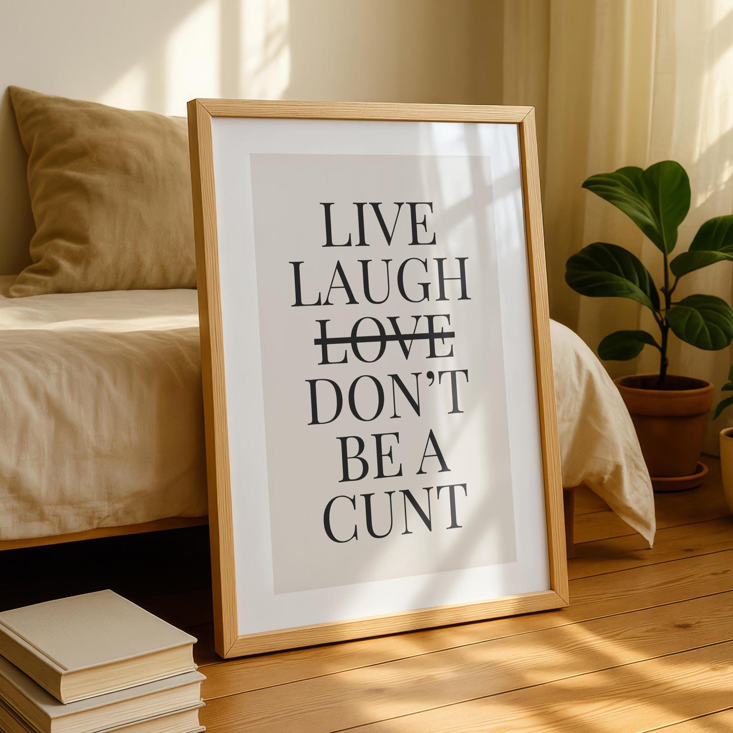 A framed print with the text "LIVE LAUGH LOVE DON'T BE A CUNT" in black on a light background, leaning against a bed with a beige duvet and pillow.