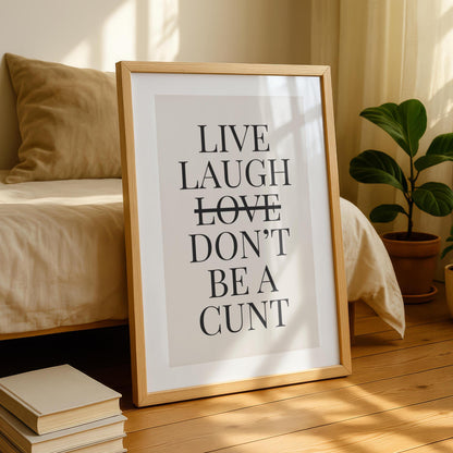 A framed print with the text "LIVE LAUGH LOVE DON'T BE A CUNT" in black on a light background, leaning against a bed with a beige duvet and pillow.