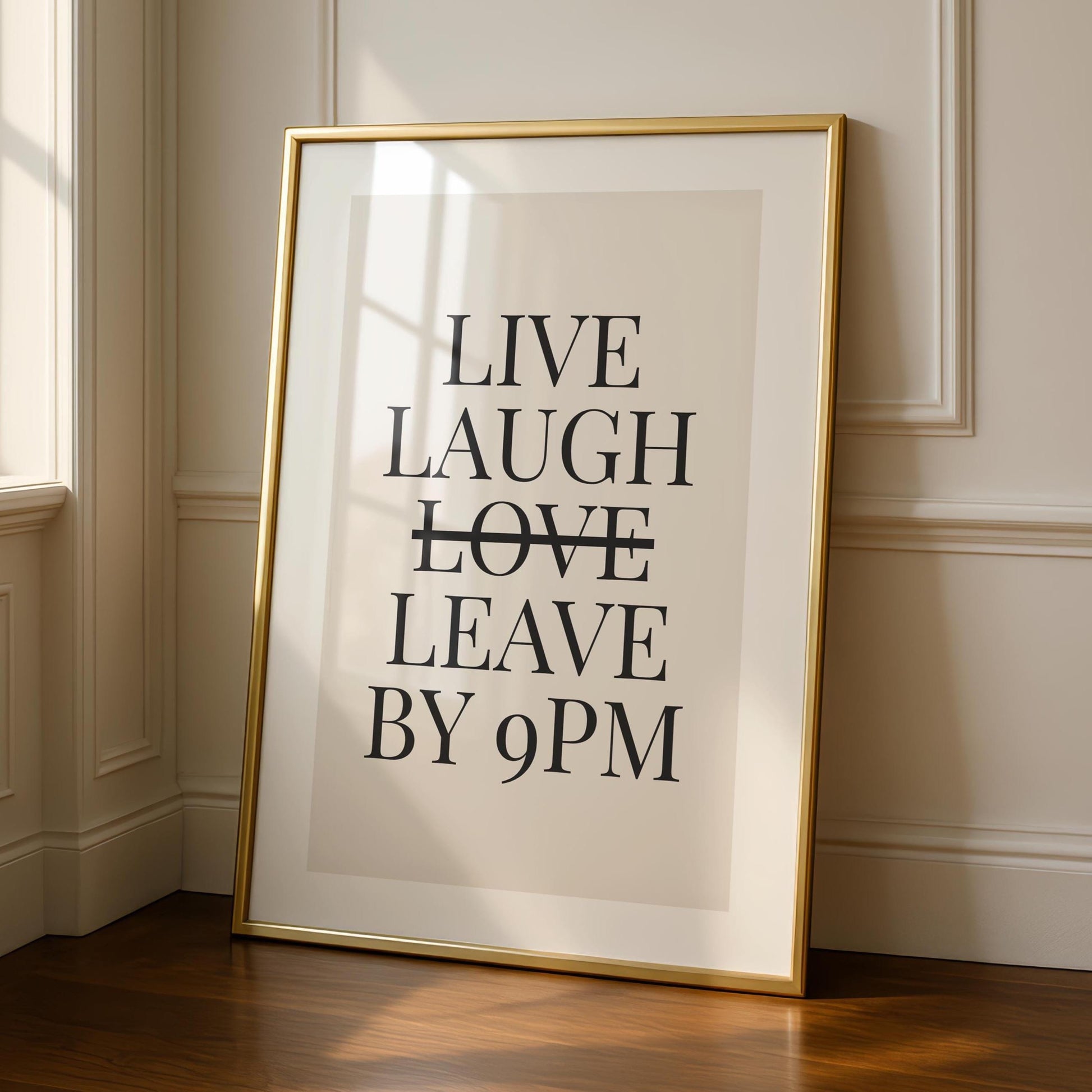A framed print with a gold border leans against a light beige wall, displaying the text "LIVE LAUGH LEAVE BY 9PM" in black sans-serif font, with a horizontal line through "LAUGH."