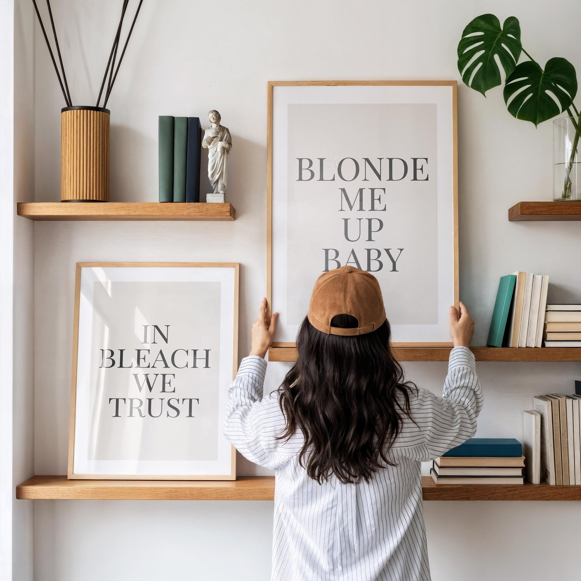 A person in a striped shirt and brown baseball cap adjusts a framed print that reads "BLONDE ME UP BABY," with another print "IN BLEACH WE TRUST" below it, on a white wall with wooden shelves.