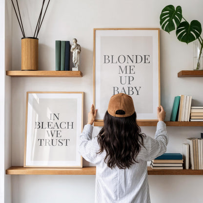 A person in a striped shirt and brown baseball cap adjusts a framed print that reads "BLONDE ME UP BABY," with another print "IN BLEACH WE TRUST" below it, on a white wall with wooden shelves.