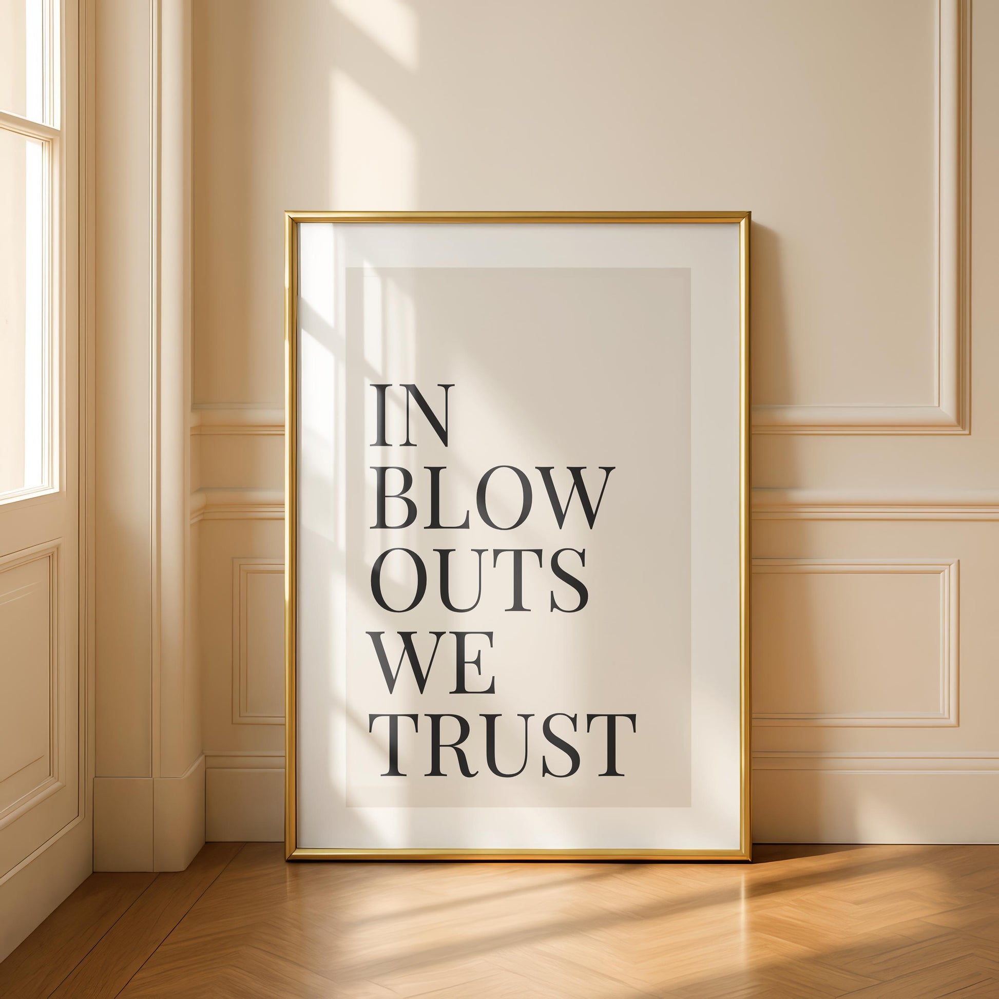 A framed print with a gold border reads "IN BLOW OUTS WE TRUST" in black text on a cream background, leaning against a white wall and wooden floor.