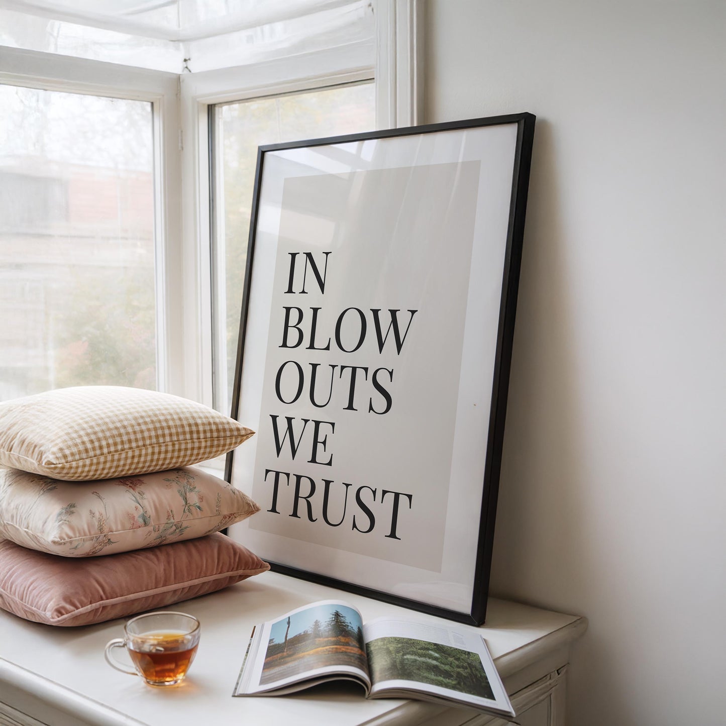 A framed print with a black border reads "IN BLOW OUTS WE TRUST" in black text on a cream background, leaning against a window with pillows and a teacup nearby.