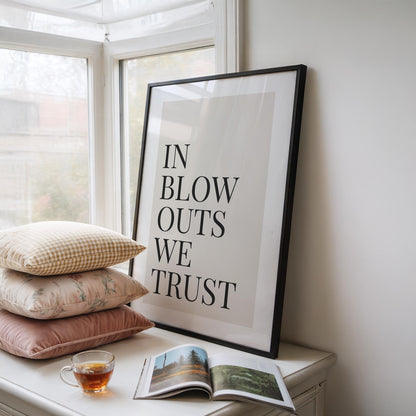A framed print with a black border reads "IN BLOW OUTS WE TRUST" in black text on a cream background, leaning against a window with pillows and a teacup nearby.