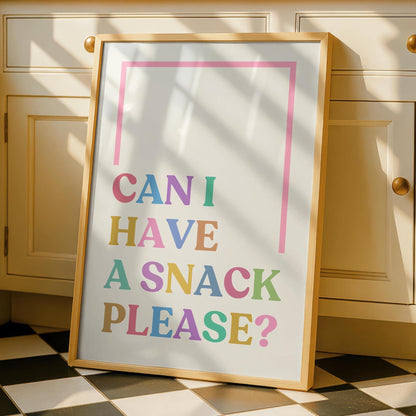 A framed print with "CAN I HAVE A SNACK PLEASE?" in colorful letters, leaning against a cream-colored kitchen cabinet.