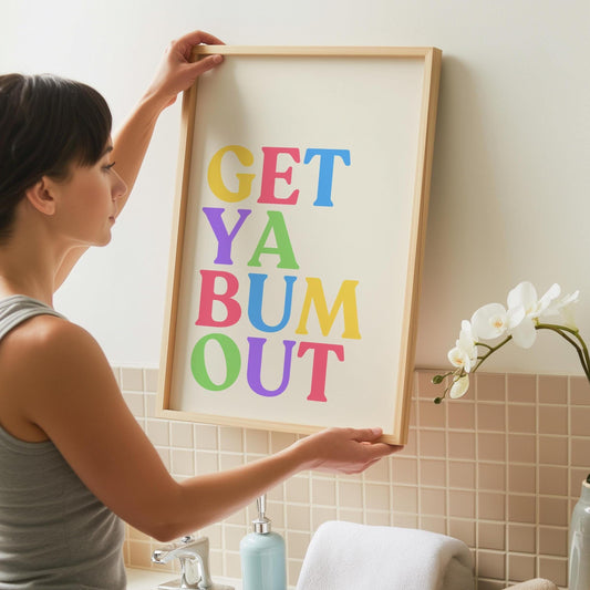 A person hangs a framed print with the words "GET YA BUM OUT" in colorful block letters on a cream background, above a sink with a light blue soap dispenser.