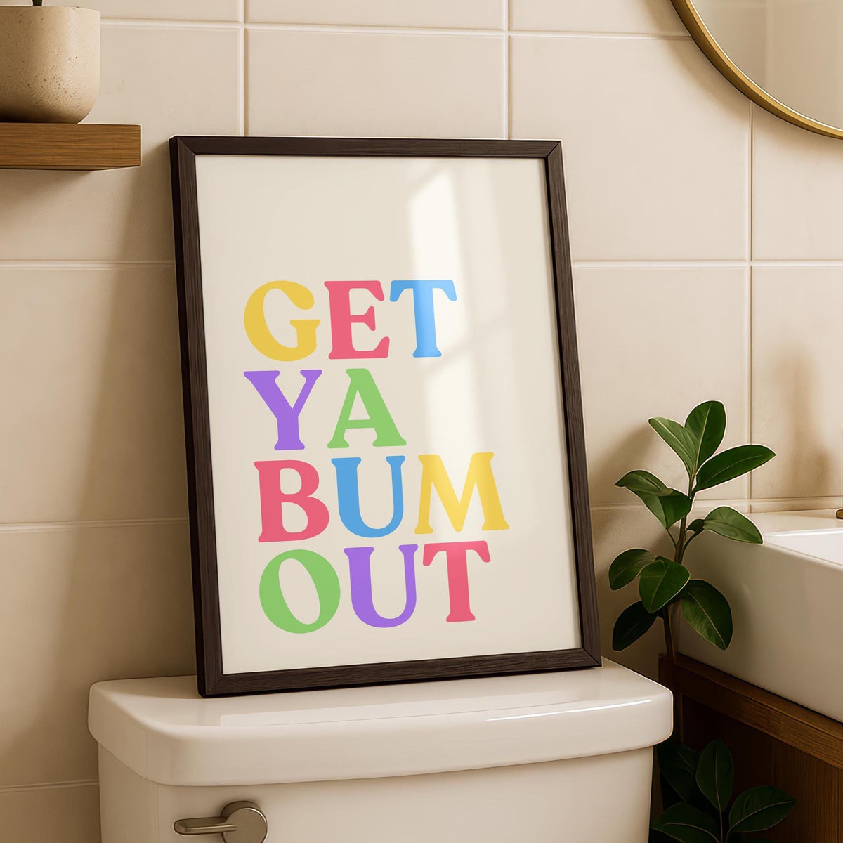 A framed print with "GET YA BUM OUT" in colorful block letters on a cream background, leaning against the wall on a toilet tank.