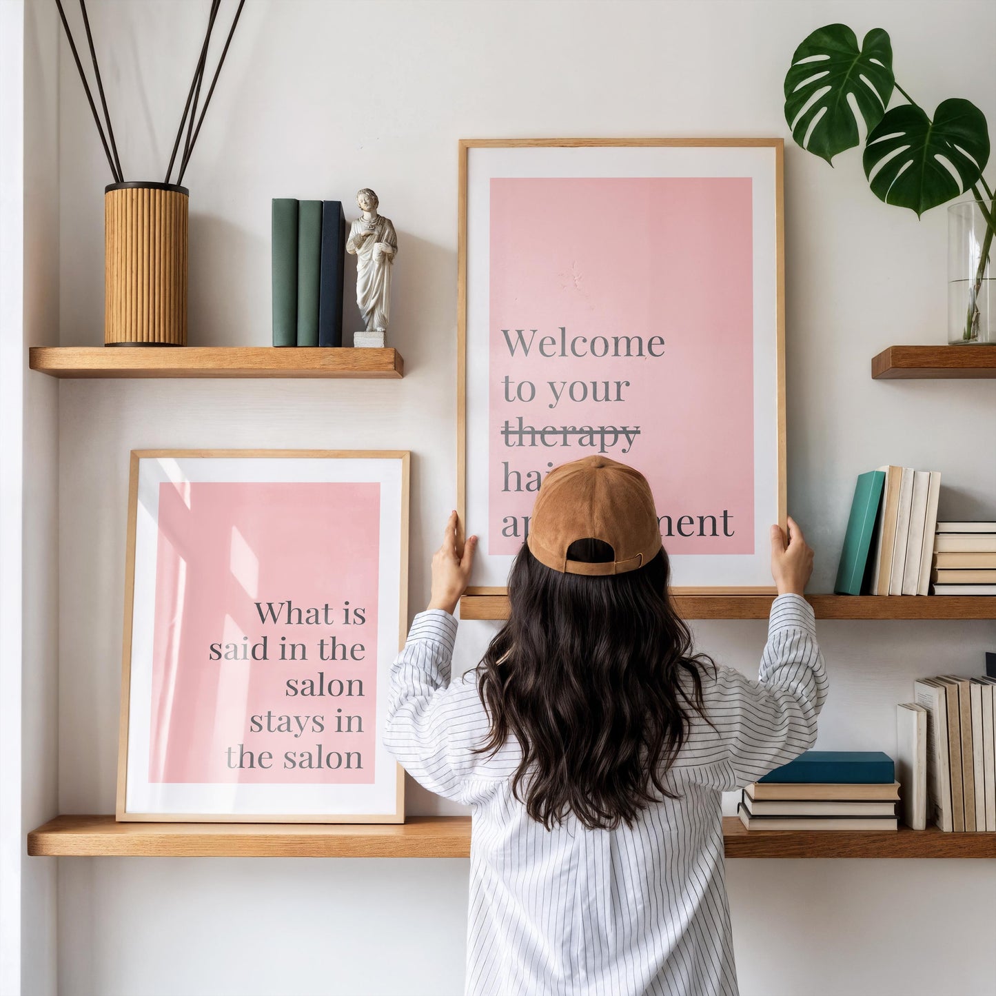 A person with long dark hair and a brown baseball cap holds a framed pink print that reads "Welcome to your therapy hair appointment" between two wooden shelves.