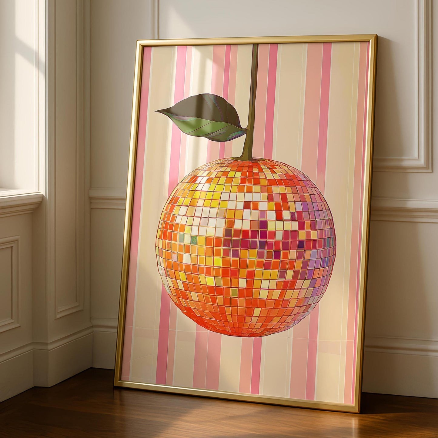 A gold-framed print of an orange disco ball with a green leaf, on a pink and beige striped background, leaning against a white wall and dark wood floor.