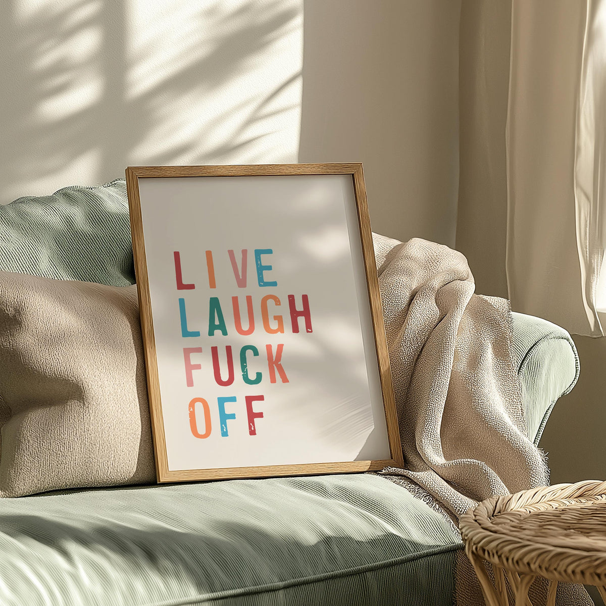A framed print with "LIVE LAUGH FUCK OFF" in colorful letters, resting on a light green sofa with a beige throw blanket, dappled with sunlight.