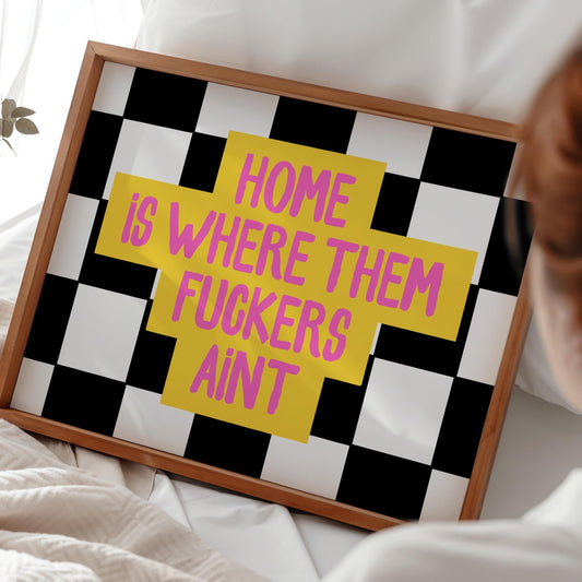 A framed print with a black and white checkerboard background and a yellow cross with the pink text "HOME IS WHERE THEM FUCKERS AINT" is held by a person in a white shirt.
