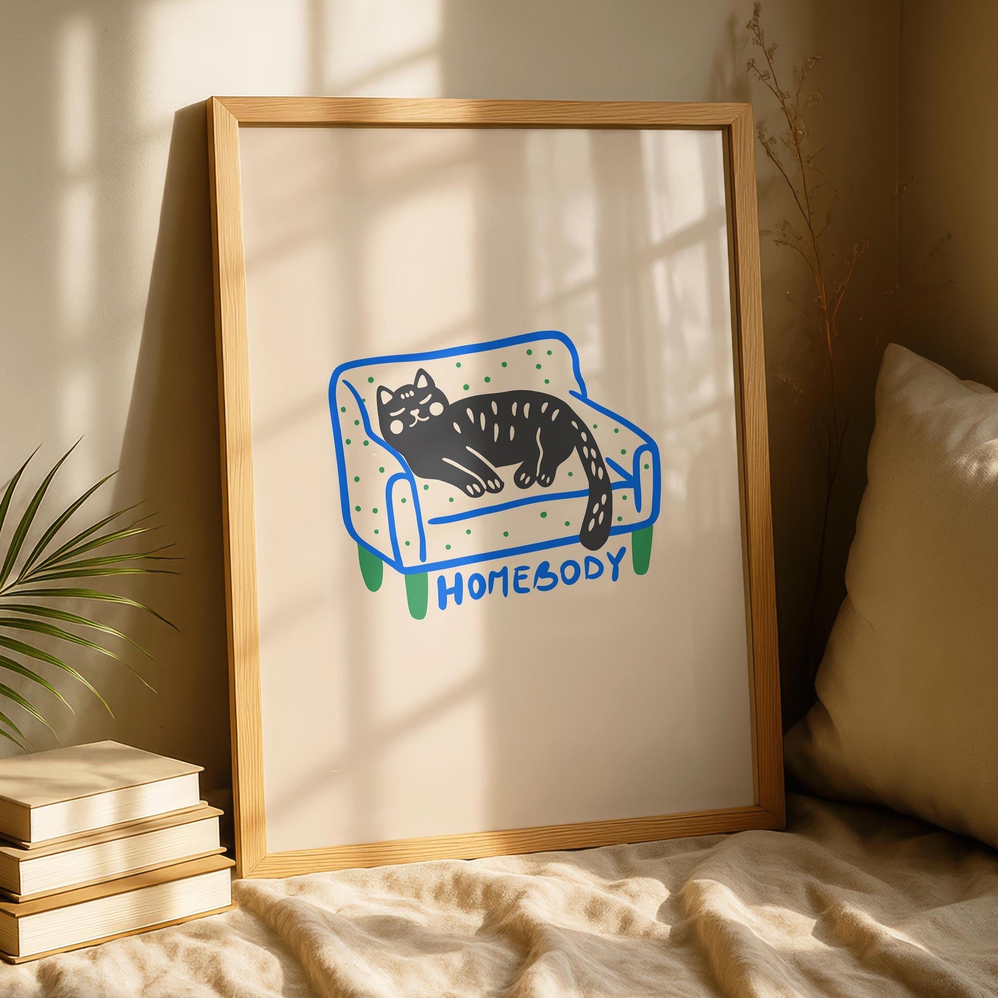 A framed print of a black cat with green eyes and white spots, sleeping on a blue and green polka-dotted couch with "HOMEBODY" written below, leaning against a wall next to a stack of books and a plant.