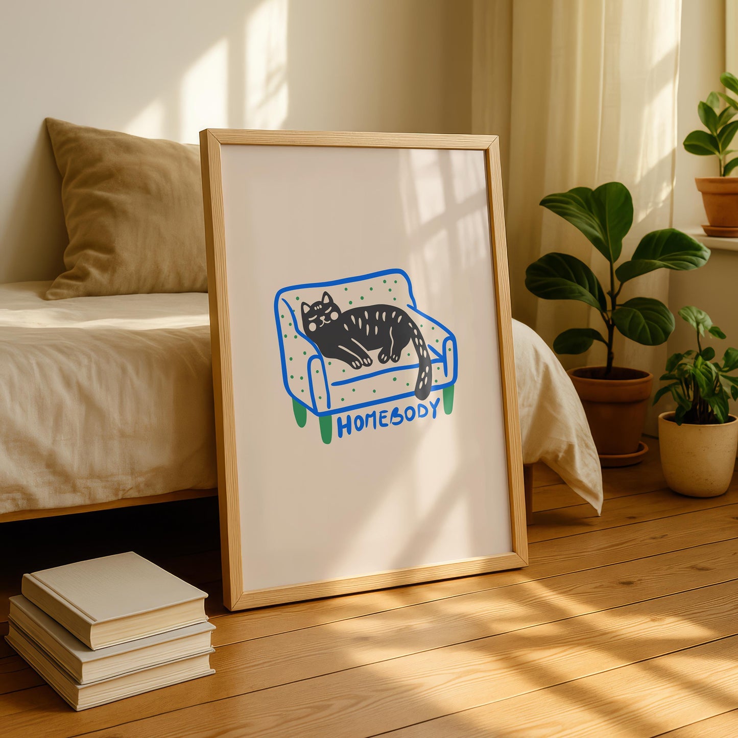 A framed print of a black cat with green eyes and white spots, sleeping on a blue and green polka-dotted couch with "HOMEBODY" written below, leaning on a wooden floor next to a bed and potted plants.