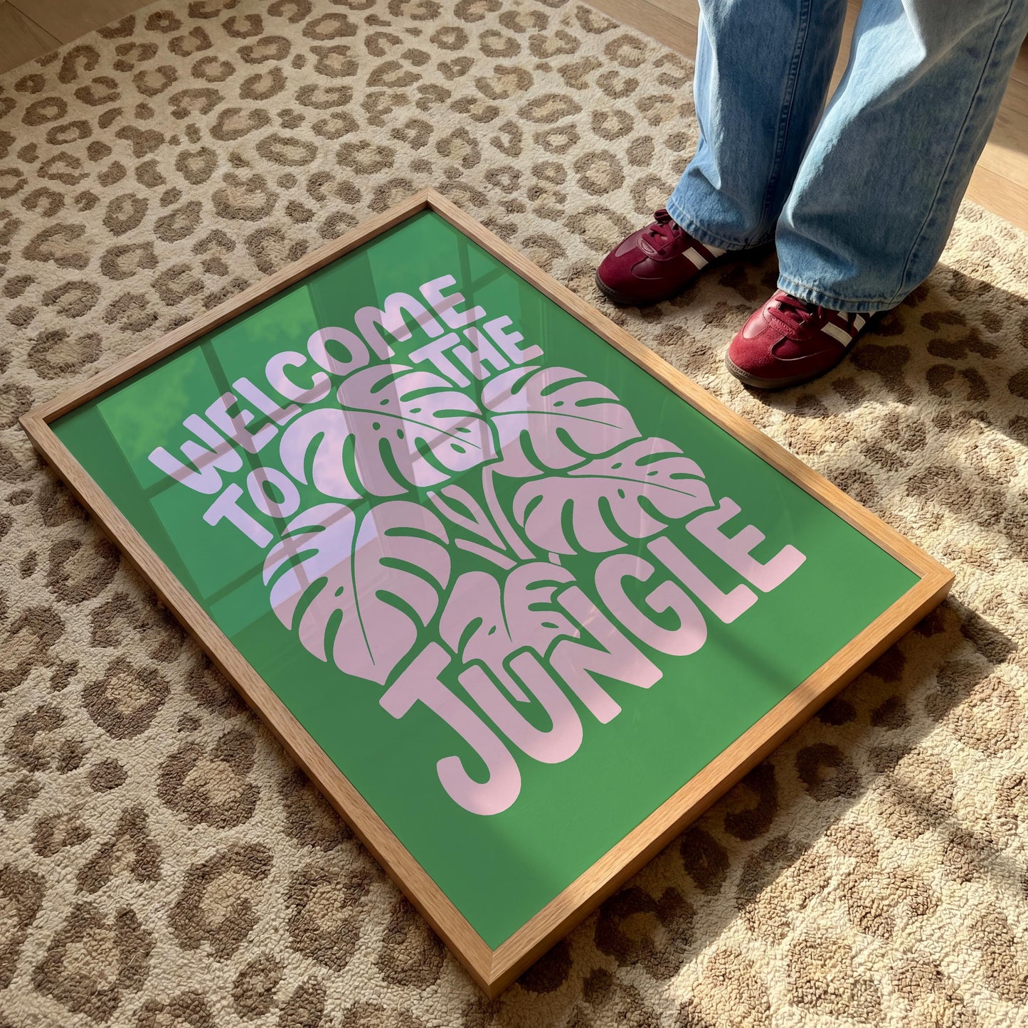 A framed green print with pink monstera leaves and the text "Welcome to the Jungle" rests on a brown leopard-print rug, with a person's blue-jeaned legs and red shoes visible at the top.