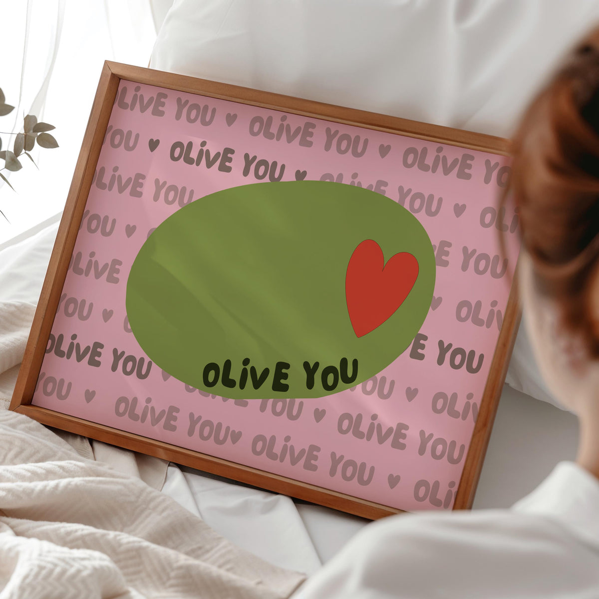 A framed print with a light pink background, a green olive with a red heart, and the words "OLIVE YOU" repeated, rests on a white bed.