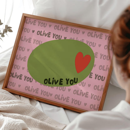 A framed print with a light pink background, a green olive with a red heart, and the words "OLIVE YOU" repeated, rests on a white bed.