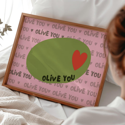 A framed print with a light pink background, a green olive with a red heart, and the words "OLIVE YOU" repeated, rests on a white bed.