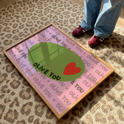 A framed print with a light pink background, a green olive with a red heart, and the words "OLIVE YOU" repeated, lies on a leopard print rug with a person standing nearby.