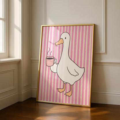 A framed print of a white goose holding a pink coffee cup with steam, on a pink and white striped background, leaning against a white wall and wooden floor.