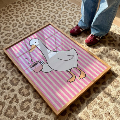 A framed print of a white goose holding a pink coffee cup with steam, on a pink and white striped background, lying on a leopard print rug with a person's feet visible.