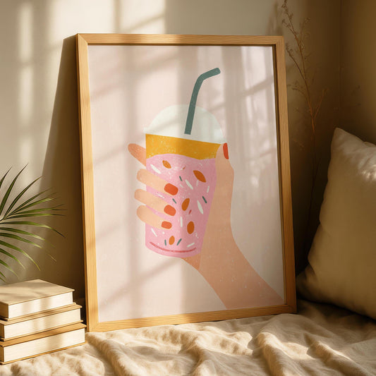 A light wood-framed print of a hand holding a pink drink with orange topping and colorful sprinkles, on a light pink background, resting on a beige bed next to a plant.