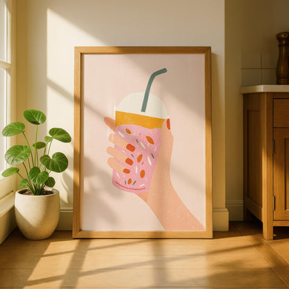 A light wood-framed print of a hand holding a pink drink with orange topping and colorful sprinkles, on a light pink background, leaning against a white wall next to a potted plant.